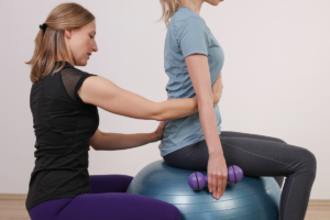 Das Bild zeigt eine Frau bei der Haltungskorrektur einer auf einem Sitzball sitzenden Frau.