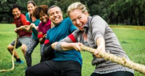 Das Foto zeigt eine Gruppe von lachenden Menschen beim gemeinsamen Tauziehen als Sinnbild für die positiven Auswirkungen durch betriebliches Gesundheitsmanagement