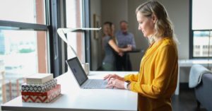 Das Foto zeigt eine Frau mit Laptop an einem Stehtisch als Sinnbild für Bewegung am Arbeitsplatz