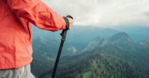 Das Bild zeigt einen Menschen mit Windjacke und Nordic-Walking Stock auf einem Berggipfel als Sinnbild für die Kombination von Bewegung und Entspannung im Rahmen von Aktivreisen und Präventionsreisen zur persönlichen Gesunderhaltung
