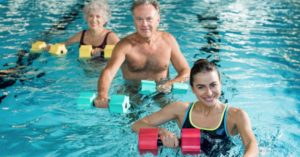 Das Bild zeigt lächelnde Menschen im hüfttiefen Wasser eines Schwimmbads beim gemeinsam Training als Sinnbild für das vielseitige Bewegungsangebot bei Aquafitness und Wassergymnastik
