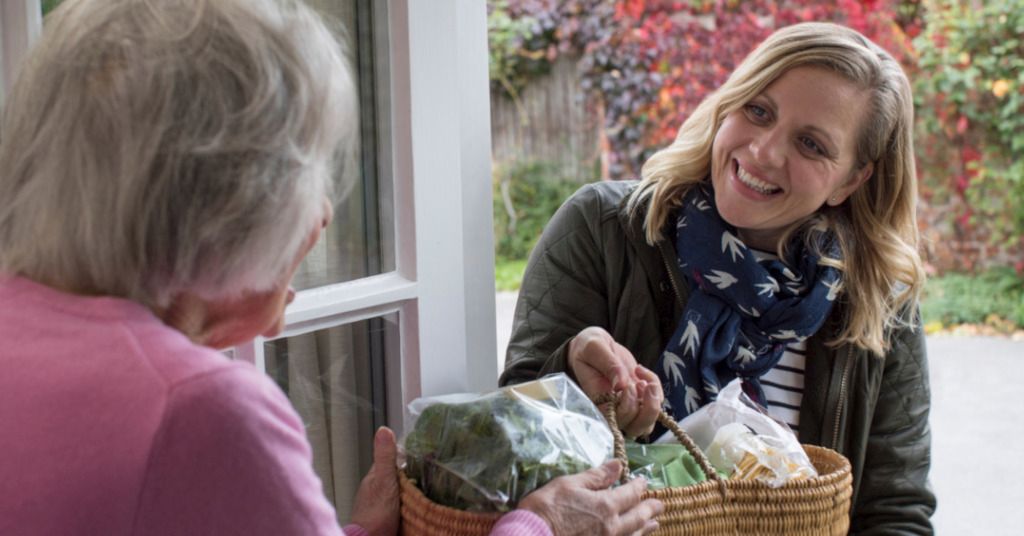 Das Bild zeigt eine Seniorin die an der Haustür von einer Frau einen Einkaufskorb mit Lebensmitteln überreicht bekommt als Sinnbild für Dienstleistungen die beschwerliche Aufgaben im Alltag erleichtern