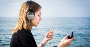 Das Foto zeigt eine Frau mit Kopfhörern und geschlossenen Augen am Meer als Sinnbild für die Entspannung durch Phantasiereisen