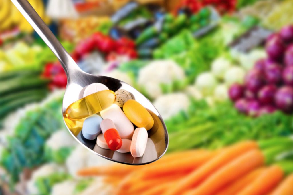 Das Foto zeigt einen Löffel mit Nahrungsergänzungsmitteln in Pillen- und Tablettenform vor vielen Gemüsen im Hintergrund als Sinnbild für den Beitrag solcher Stoffe zur richtigen Ernährung gegen Knieschmerzen