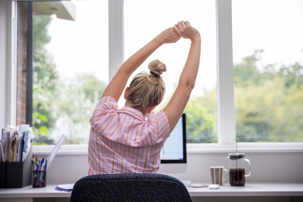 Das Bild zeigt eine Frau beim Strecken am Computerarbeitsplatz um auf die Bedeutung von Bewegung im Homeoffice hinzuweisen.