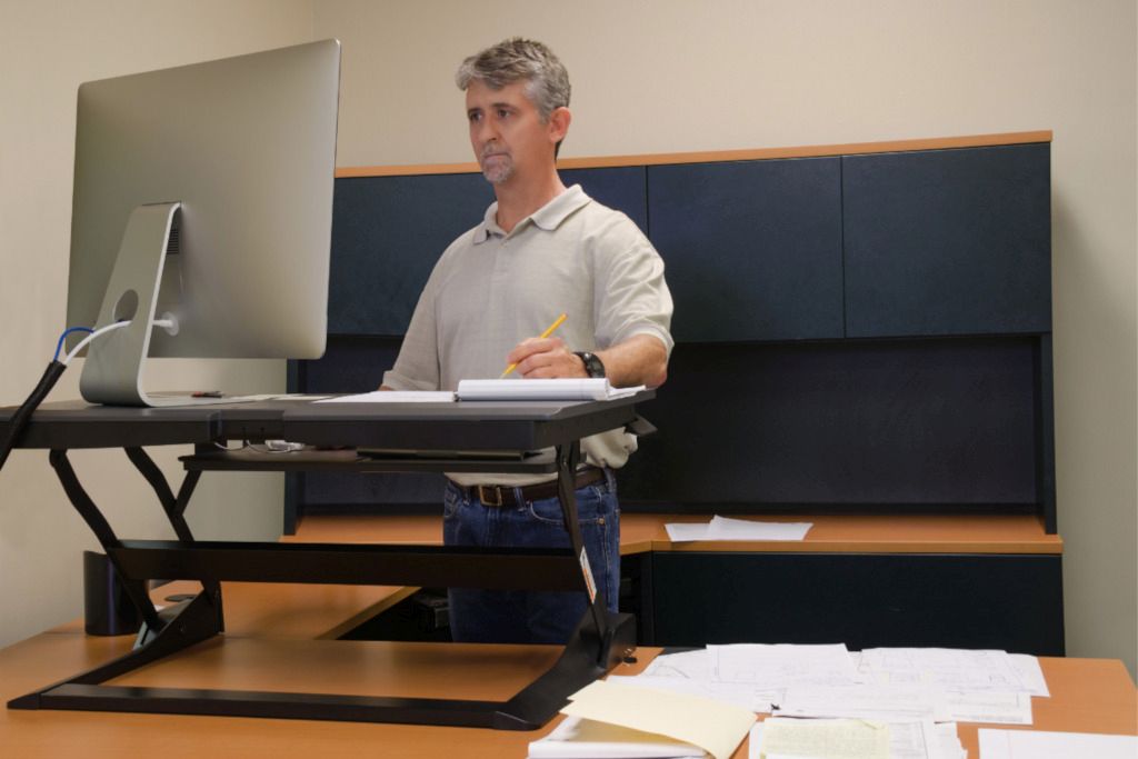 Das Bild zeigt einen Mann an einem höhenverstellbaren Schreibtischaufsatz um auf gute ergonomische Steharbeitsplätze im Homeoffice hinzuweisen.