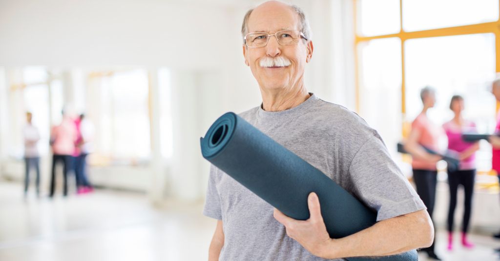 Das Foto zeigt einen strahlenden Senior mit Gymnastikmatte um den Spaß und Wert von Übungen für Senioren in einem Kurs anzudeuten