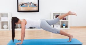 Das Foto zeigt eine Frau beim Pilates auf einer Matte im Wohnzimmer vor dem Fernseher auf dem ein angeleiteter Pilates-Kurs zu sehen ist.