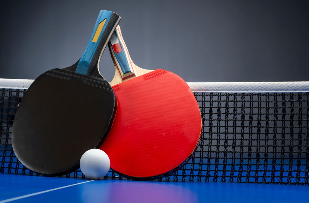 Das Foto zeigt zwei typische Tischtennisschläger in Rot und Schwarz und einen weißen Ball als Ausstattung um Tischtennis spielen zu können