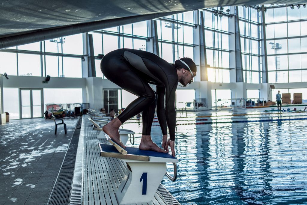 Das Bild zeigt einen Schwimmer im Ganzkörperanzug auf dem Startblock im Schwimmbad