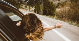 Das Foto zeigt eine Frau, die den Kopf bei einer langen Fahrt durch die Berge aus dem Autofenster hält als Sinnbild für das Thema Verspannungen in Folge langer Reisen mit dem Auto