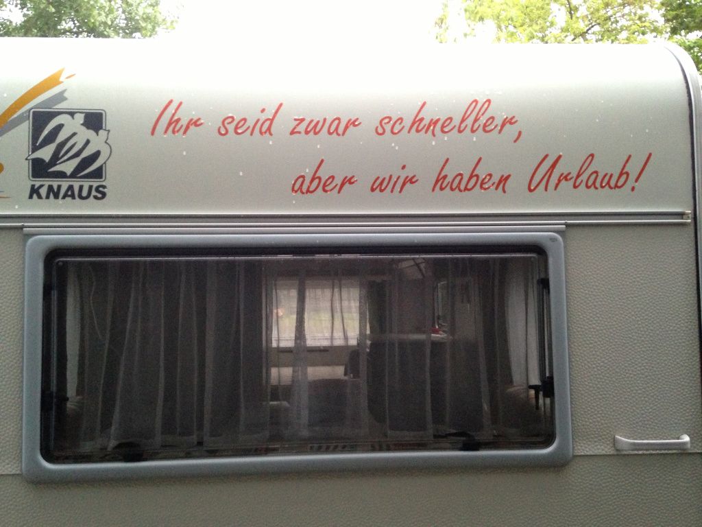 Das Foto zeigt das Heck eines Wohnwagens mit dem Spruch "Ihr seid zwar schneller, aber wir haben Urlaub" als Sinnbild für eine Urlaubsreise mit dem Auto ohne Verspannungen