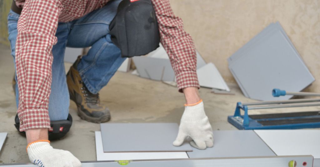 Das Bild zeigt einen Handwerker mit Kniepolstern beim Fliesenlegen als Sinnbild für den Einsatz schützender Arbeitsmittel wie Knieschoner oder Kniekissen