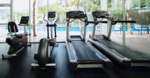 Das Bild zeigt eine Reihe unterschiedlicher Cardio Fitnessgeräte in einem Raum mit großer Glasscheibe und einem sichtbaren Außenpool als Sinnbild für ein ansprechendes und wirksames Training zuhause mit der richtigen Ausstattung