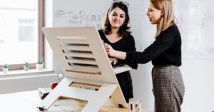 Das Bild zeigt zwei Frauen bei der Arbeit im Büro an einem modernen und ergonomischen Schreibtischaufsatz als Sinnbild für die entlastende Nutzung höhenverstellbarer Schreibtische und Aufsätze
