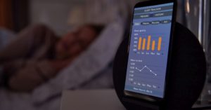 Das Bild zeigt ein Smartphone mit einem Programm zur Erfassung der Schlafdaten neben einem schlafenden Menschen als Sinnbild für die Möglichkeit, mit dem richtigen Fitness Tracker den Schlaf zu überwachen