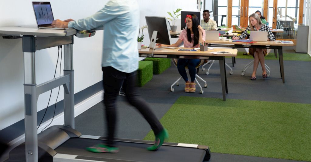Das Bild zeigt einen Mann auf einem Laufband bei der Arbeit am Laptop in einem Großraumbüro vor einer Reihe von Kollegen an ihren Schreibtischen als Sinnbild für moderne Fitnessgeräte die Sport und Bewegung am Arbeitsplatz ermöglichen