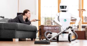 Das Bild zeigt eine Frau bei der Pause auf dem Sofa während ein Roboter die Hausarbeit erledigt: So kannst du Smart-Home Systeme von Alexa bis zum Mähroboter zu deiner Entlastung nutzen