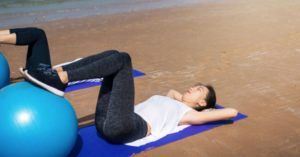 Das Bild zeigt eine Frau beim Rückentraining auf einer Yogamatte am Strand als Sinnbild für die tollen Möglichkeiten ein Online-Training für die Steigerung des Wohlbefindens zu nutzen