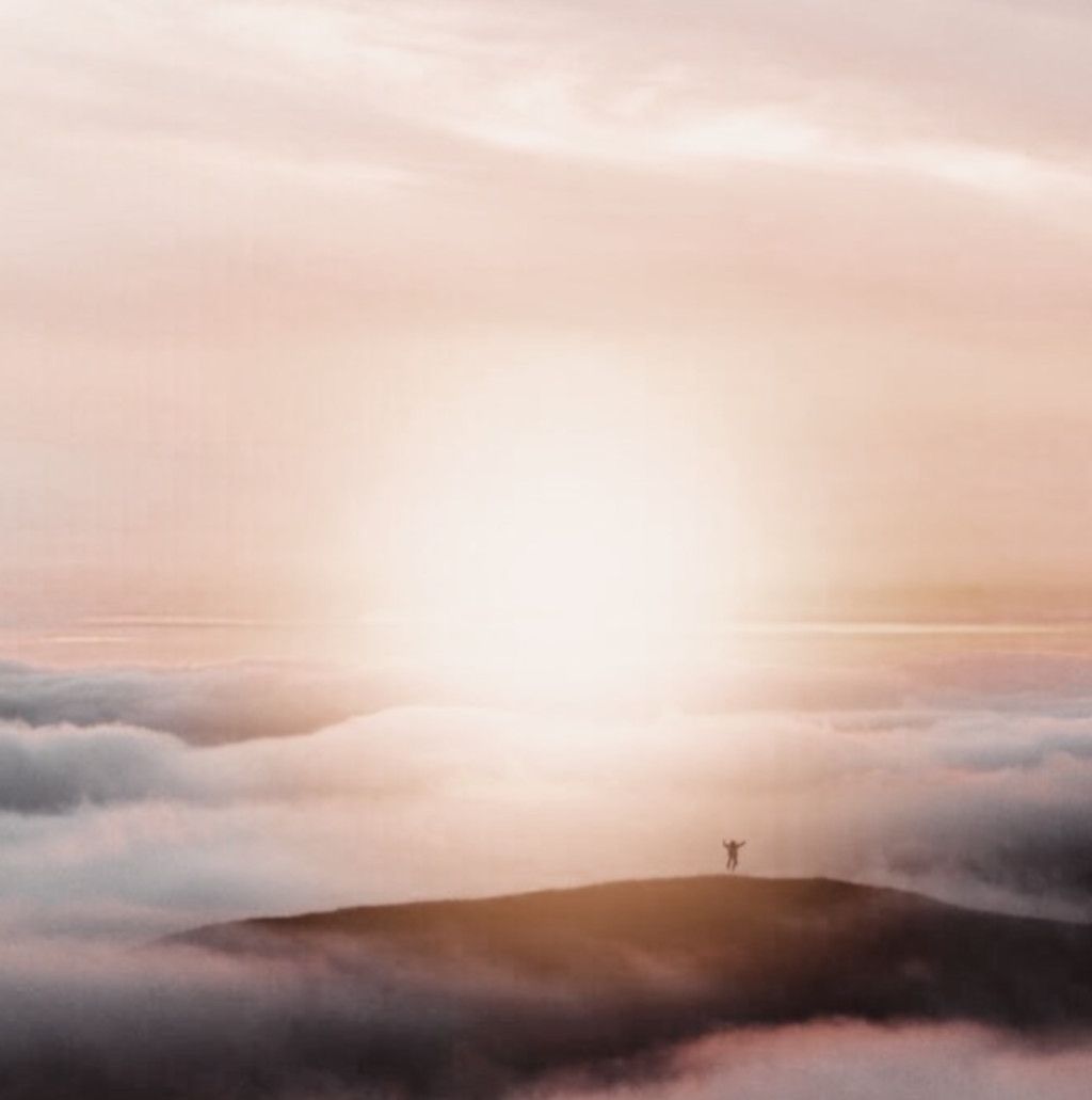 Das Bild zeigt einen Menschen auf einer Bergspitze im Sonnenlicht oberhalb der geschlossenen Wolkendecke als Sinnbild für die Säule "Optimismus" bei der Resilienz