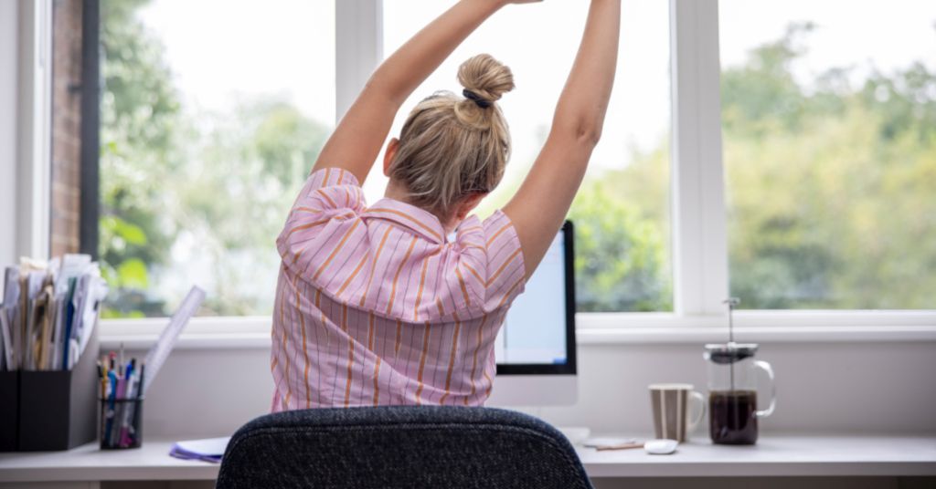 Das Bild zeigt eine Frau bei angeleiteten Dehnübungen am Schreibtisch im Büro als Sinnbild für eine aktive Pause als Teil der Gesundheitsförderung für Mitarbeitende im Betrieb
