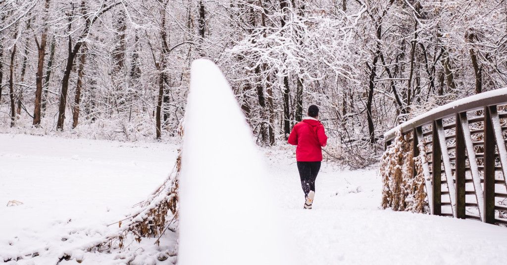 Das Bild zeigt einen Menschen beim Joggen in einer verschneiten Landschaft im Wald als Sinnbild für schönen und gesunden Sport in der Kälte