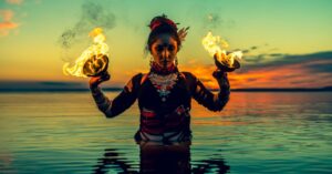 Das Bild zeigt eine schöne und stolze Frau mit zwei Feuerschalen in den Händen im Wasser stehend als Sinnbild dafür, den eigenen Körper als lebendiges Ausdrucksmittel der eigenen Weiblichkeit zu würdigen.