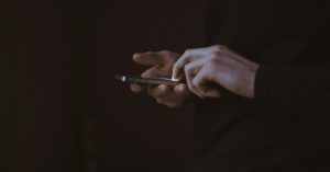 Das Bild zeigt zwei menschliche Hände bei der Nutzung eines Smartphones vor einem dunklen Hintergrund als Sinnbild für das Suchen von passenden digitalen Informationen von Menschen mit chronischer Erkrankung zur Stärkung der eigenen Gesundheitskompetenz.