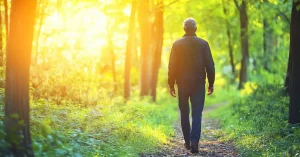 Das Bild zeigt einen Geschäftsmann beim Spaziergang im sonnigen Wald als Sinnbild dafür, bereits mit einer kurzen täglichen Meditation und Achtsamkeit wieder mehr Energie zu bekommen und Burnout vorzubeugen.