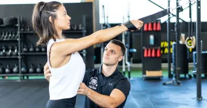 Das Bild zeigt einen CorssFit-Trainer dabei, wie er einer jungen Frau bei der Ausführung einer Übung hilft, die für die Präventation von Rückenschmerzen ist.