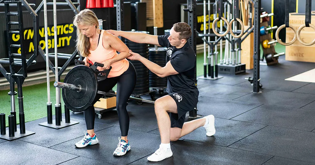 Das Bild zeigt einen CorssFit-Trainer dabei, wie er einer Frau bei der Ausführung einer Übung hilft, die für die Präventation von Rückenschmerzen ist.